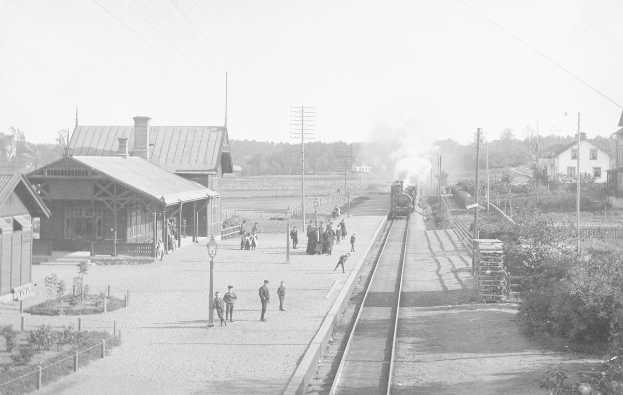 Schwarz-weiß-Foto eines Zuges, der an einem Bahnhof mit wartenden Menschen auf dem Bahnsteig hält, umgeben von Straßeninfrastruktur und Gebäuden.