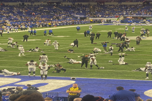 Ein Fußballspiel wird in einem Stadion gespielt, während Menschen auf dem Rasen und einige auf Stühlen im Hintergrund stehen und eine Person eine Kamera hält, was auf eine Live-Übertragung hinweist.