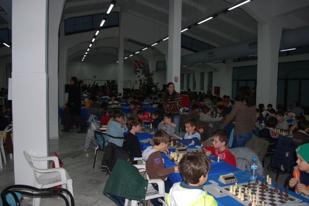 Eine Gruppe von Menschen, die um einen Tisch herum auf Stühlen sitzen und Schach spielen in einem Saal mit einer weißen Decke, montierten Leuchten und grauen Säulen.