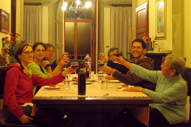 Eine Gruppe von Menschen an einem Tisch sitzend, die Weinpokale halten, mit Flaschen, Tellern, einem Foto auf der Wand und Vorhängen im Hintergrund.