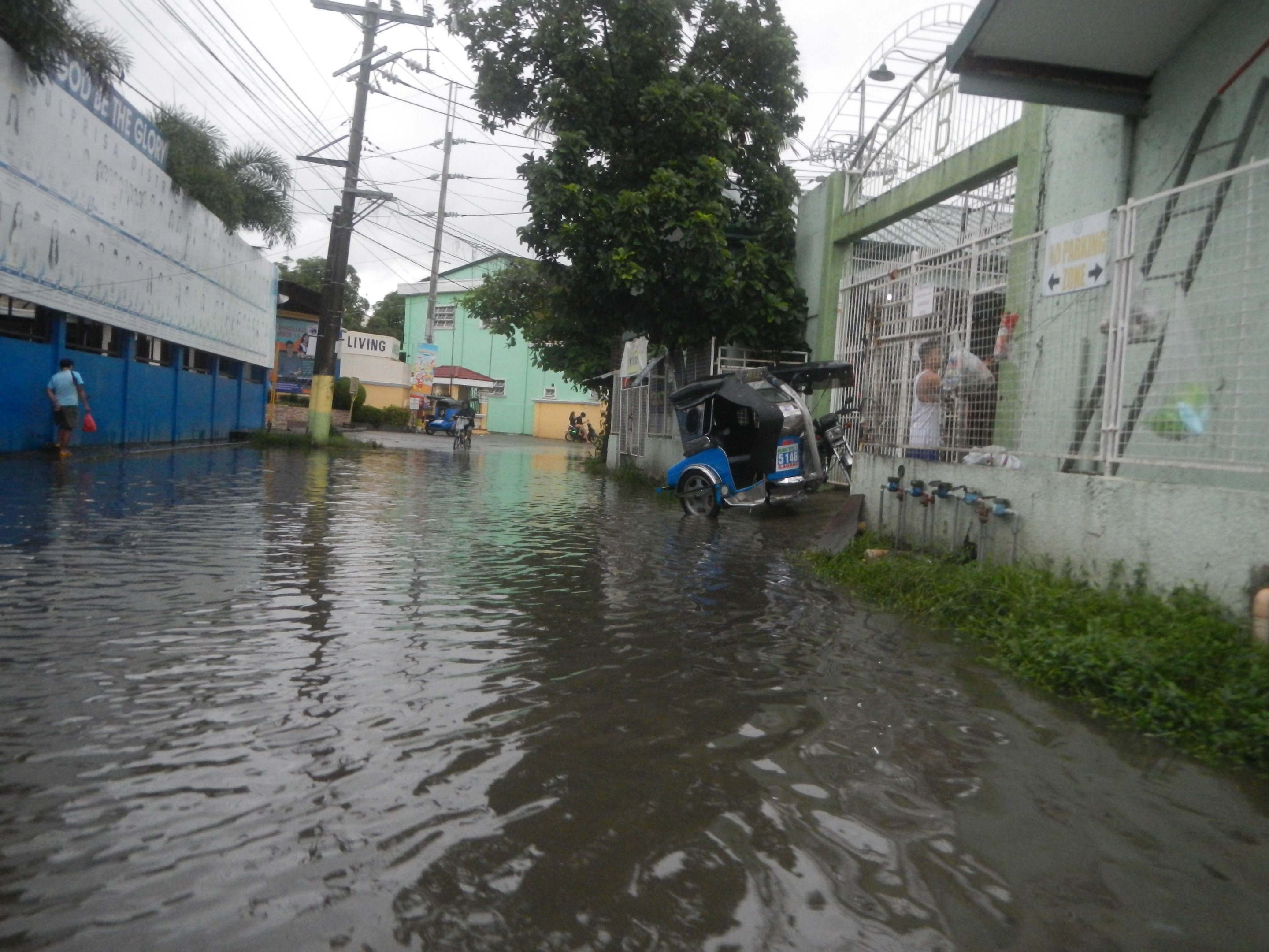 Eine überflutete Straße mit einem Motorrad in der Mitte, flankiert von Gebäuden, Bäumen und Strommasten mit Überlandleitungen und einigen Menschen und Fahrzeugen im Hintergrund unter einem sichtbaren Himmel.