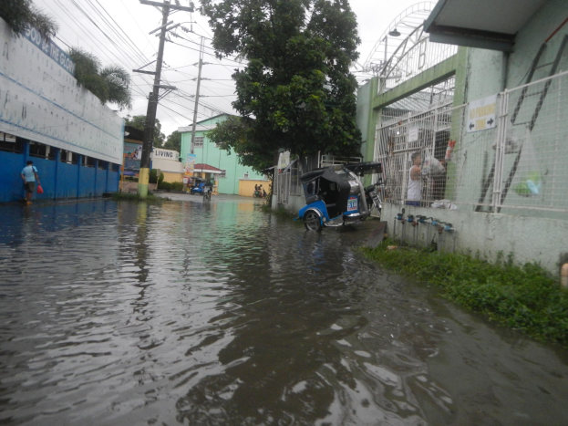 Eine überflutete Straße mit einem Motorrad in der Mitte, flankiert von Gebäuden, Bäumen und Strommasten mit Überlandleitungen und einigen Menschen und Fahrzeugen im Hintergrund unter einem sichtbaren Himmel.