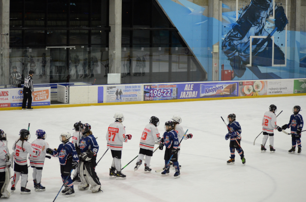 Gruppe junger Menschen beim Eisschnelllaufen auf einer Indoor-Eisfläche, die Helme tragen und Hockey-Schläger halten, mit einer Person auf der linken Seite und Bannern, Säulen und Glaswänden im Hintergrund.