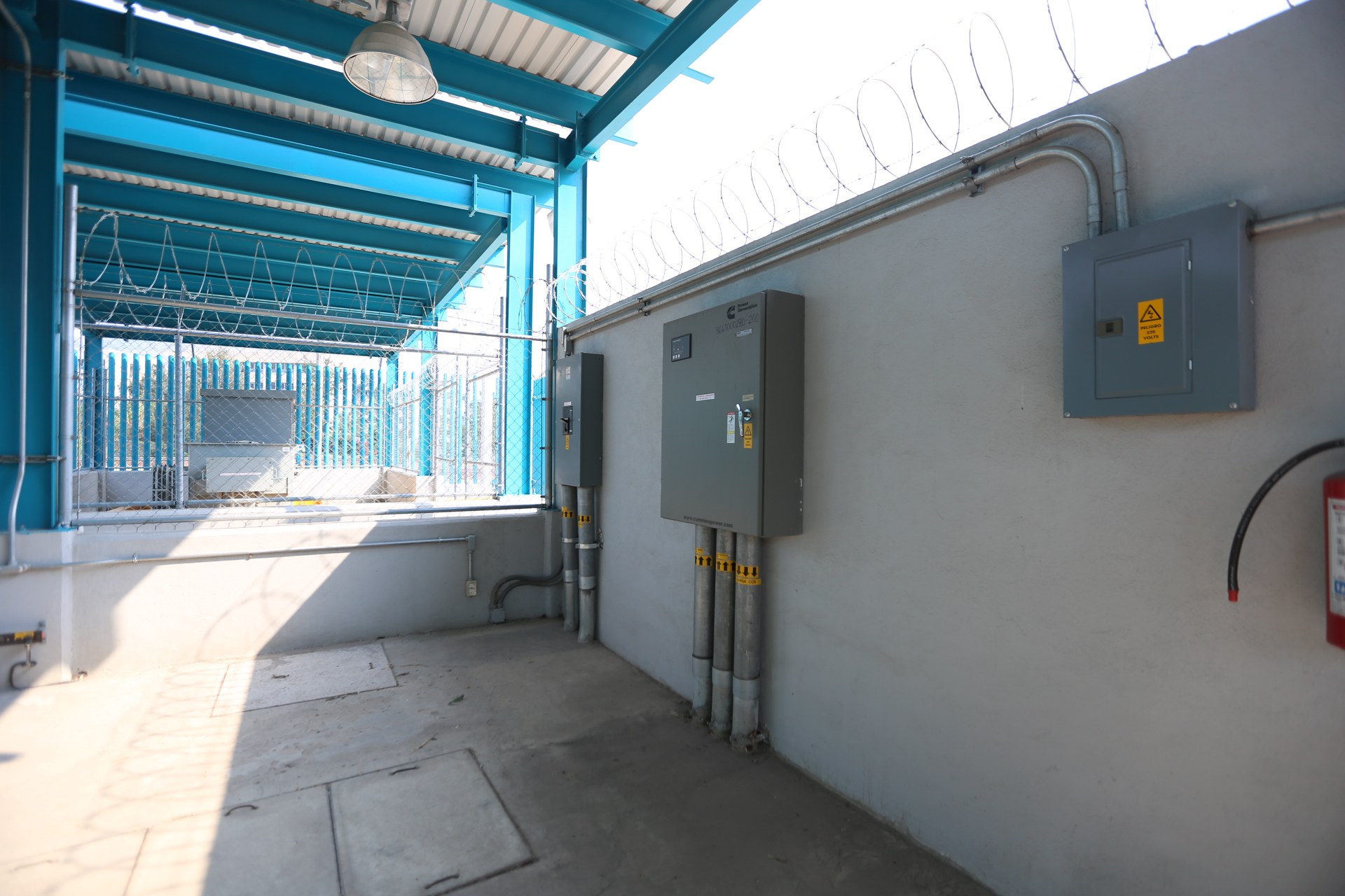 Ein Raum mit einem Feueröscher auf der rechten Seite, elektrischen Schaltkästen und Rohren an der Wand, einer Deckenleuchte und einem Zaun im Hintergrund, ähnlich einer elektrischen Umspannstation.
