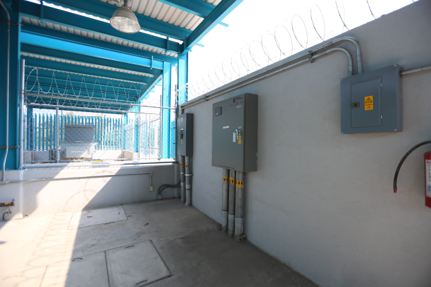 Ein Raum mit einem Feueröscher auf der rechten Seite, elektrischen Schaltkästen und Rohren an der Wand, einer Deckenleuchte und einem Zaun im Hintergrund, ähnlich einer elektrischen Umspannstation.