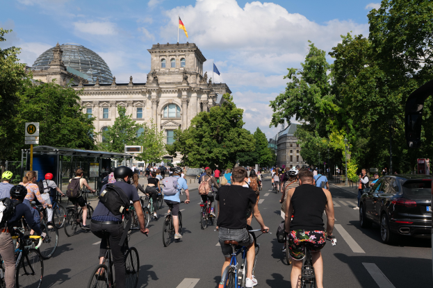 Eine Gruppe von Menschen fährt Fahrrad auf einer von Bäumen gesäumten Straße in Berlin, Deutschland, mit Gebäuden auf beiden Seiten, einem Bushaltestellen auf der rechten Seite und einer Flagge, die auf einem der Gebäude weht, unter einem bewölkten Himmel.