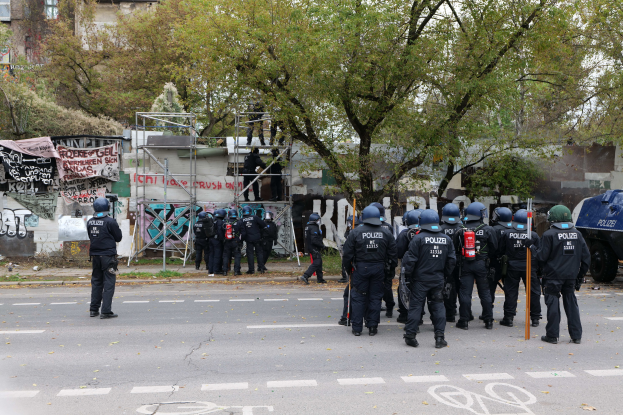 Polizeibeamte in schwarzen Uniformen und Helmen stehen vor einem Graffiti-bedeckten Gebäude, einige halten Stöcke, mit Bäumen, Gebäuden, einem klaren Himmel und einem geparkten Fahrzeug im Hintergrund.