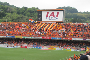 Ein Fußballspiel wird in einem Stadion mit einer großen Zuschauermenge, grünem Rasen, einem Tor, Bannern, Fahnen, einem großen Bildschirm, Bäumen und einem klaren blauen Himmel gespielt.