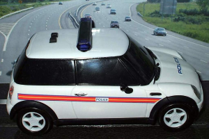 Weißer Polizeiwagen vor einer belebten Straße mit Fahrzeugen, Gras und Bäumen auf beiden Seiten, aufgenommen außerhalb einer Stadt.