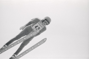 Ein Skispringer in voller Ausrüstung springt mitten in der Luft auf Skiern gegen einen klaren blauen Himmel.