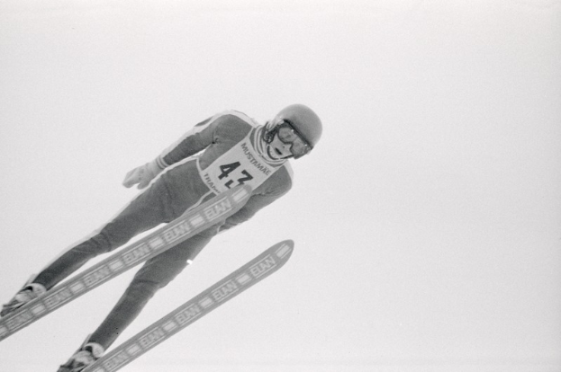 Ein Skispringer in voller Ausrüstung springt mitten in der Luft auf Skiern gegen einen klaren blauen Himmel.