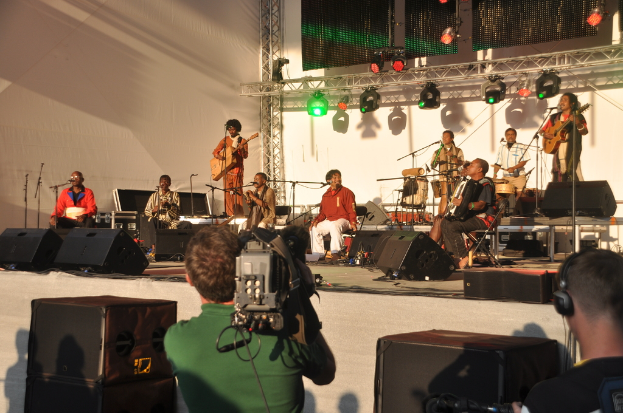 Ein musikalisches Konzert mit Musikern, die verschiedene Instrumente auf der Bühne spielen, darunter Gitarren und Trommeln, unter rot und grünen Scheinwerfern, mit Lautsprechern vorne und zwei Personen, eine filmt und die andere hört zu.