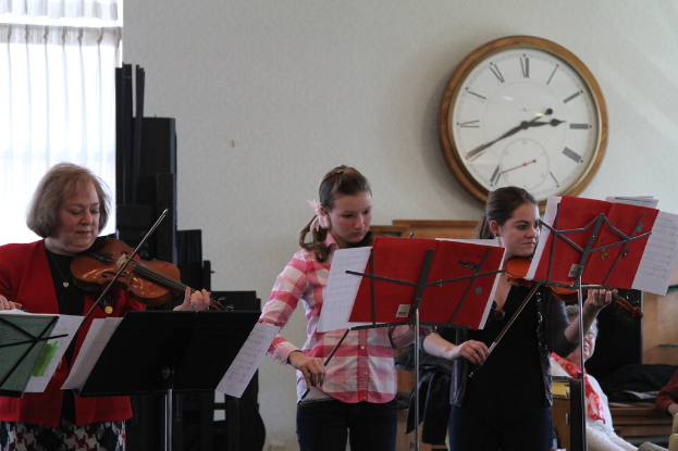 Drei Frauen auf der Bühne, die Geigen spielen, mit Notenpulten und Noten davor, während ein Zuschauer im Vordergrund sitzt und zuhört; eine Uhr hängt an der weißen Wand im Hintergrund, und ein weißer Vorhang ist links sichtbar.