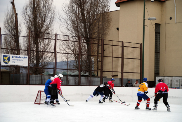 Personen beim Eishockey auf einer Eisfläche mit Gebäuden, Bäumen, einer Straßenlaterne, einem Namensschild und Zäunen im Hintergrund bei klarem Himmel.