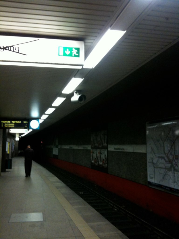 Eine Person steht auf einem Bahnsteig mit einem Namen auf der Wand, Lampen und einer Kamera auf dem Dach, mit einem Zug auf den Schienen rechts.