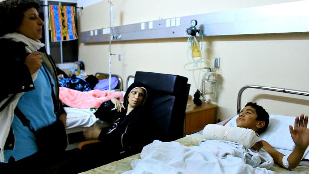 Ein Krankenhauszimmer mit mehreren Betten, auf denen Patienten liegen, Infusionsflaschen auf Ständern zwischen den Betten und eine Frau mit einer Tasche in der Nähe eines der Patienten.