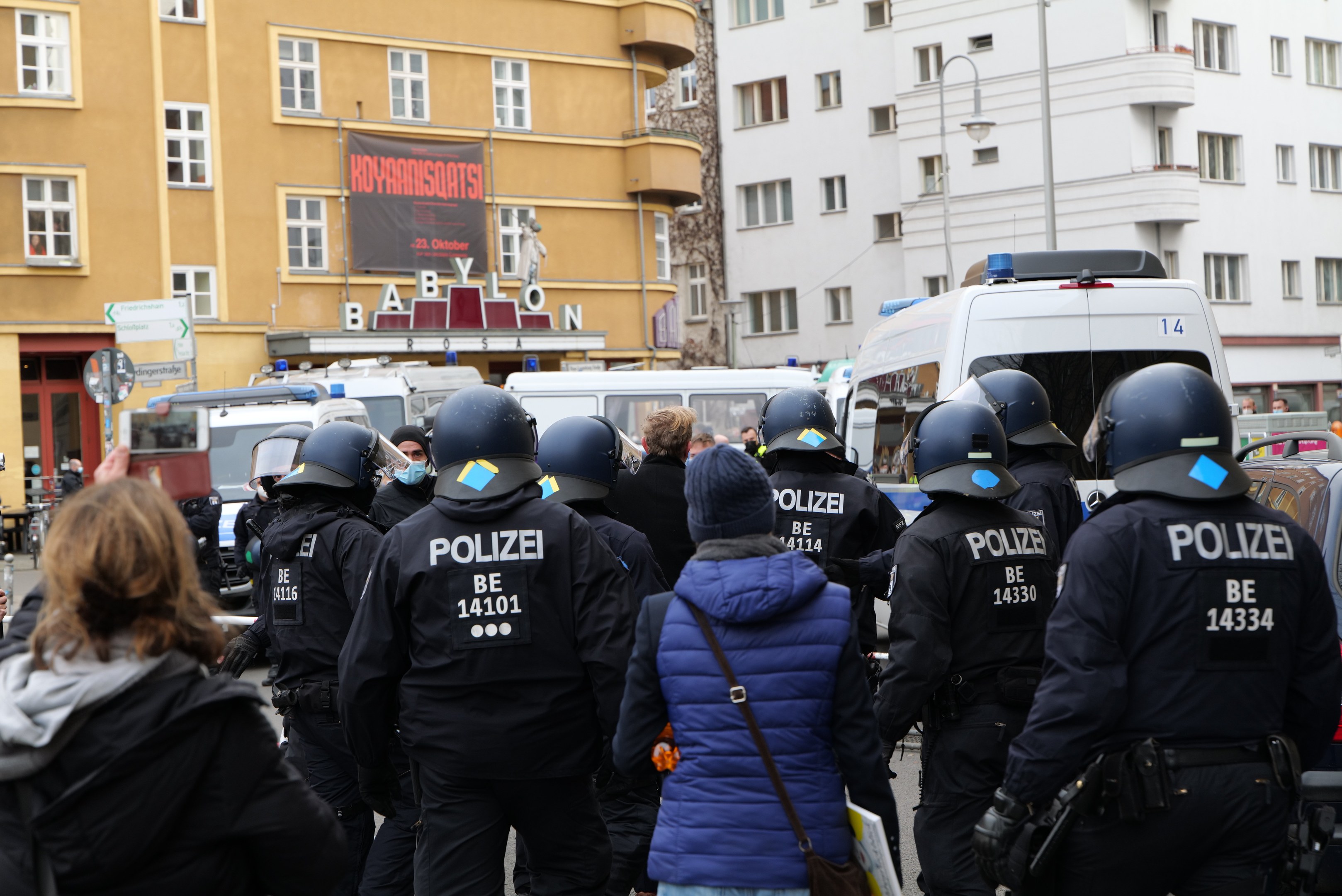 Polizeibeamte in Uniform vor einer Menge bei einer Demonstration in Berlin, mit Fahrzeugen, Gebäuden und einer Kamera im Hintergrund.