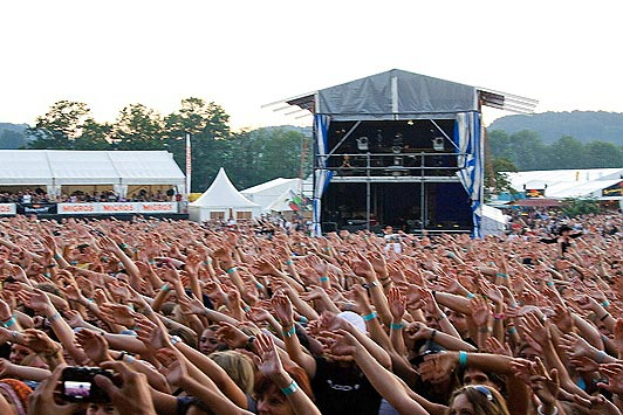 Eine große Menschenmenge auf einem Musikfestival mit erhobenen Händen, einige halten Mobiltelefone, vor einer Bühne, weißen Zelten und Bäumen im Hintergrund unter einem klaren blauen Himmel.