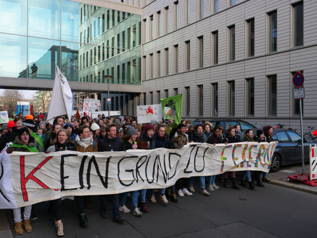 Eine Gruppe von Menschen, die auf einer Straße stehen und ein Transparent mit der Aufschrift "Kein Grind Lo Feuer" tragen, vor dem Hintergrund von Gebäuden, Bäumen, Polen, Schildern, Fahrzeugen und einem klaren blauen Himmel.