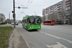 Ein grüner Bus fährt auf einer von hohen Gebäuden gesäumten Straße, mit Gras, Strommasten und Drähten auf der linken Seite und Bäumen und einem klaren blauen Himmel im Hintergrund.