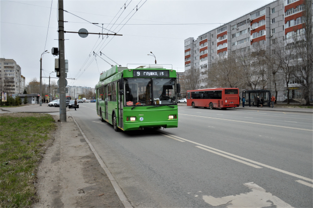 Ein grüner Bus fährt auf einer von hohen Gebäuden gesäumten Straße, mit Gras, Strommasten und Drähten auf der linken Seite und Bäumen und einem klaren blauen Himmel im Hintergrund.