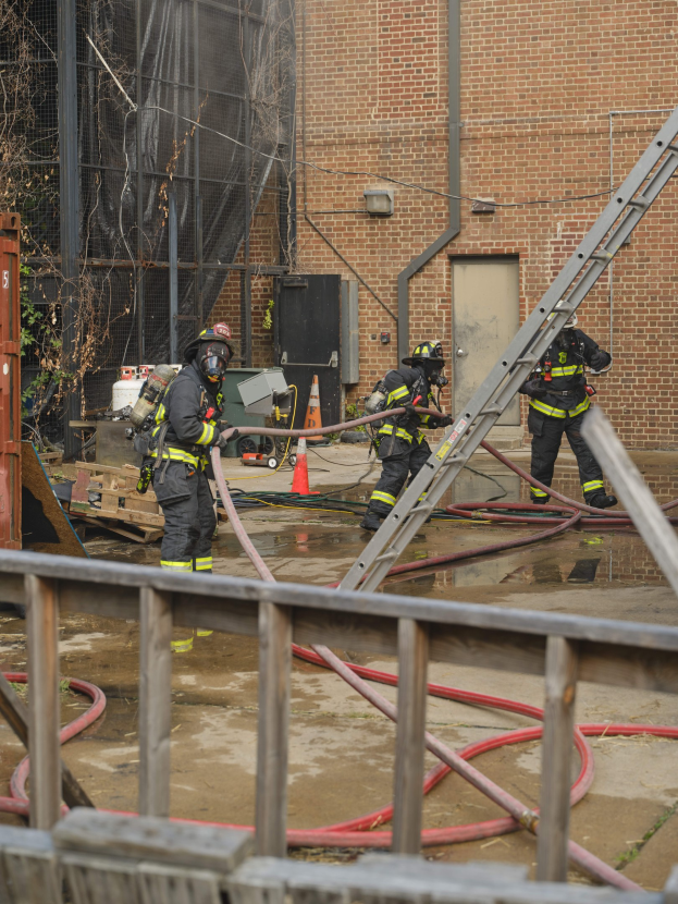 Feuerwehrleute in Helmen arbeiten daran, ein Gebäude Feuer zu löschen, umgeben von Equipment und einem Metallzaun.