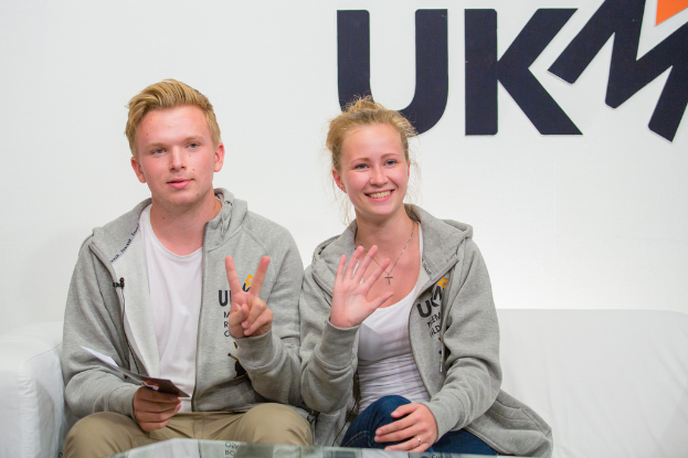 Ein Paar in passenden Jacken auf einem Sofa mit 'UKM' im Hintergrund.