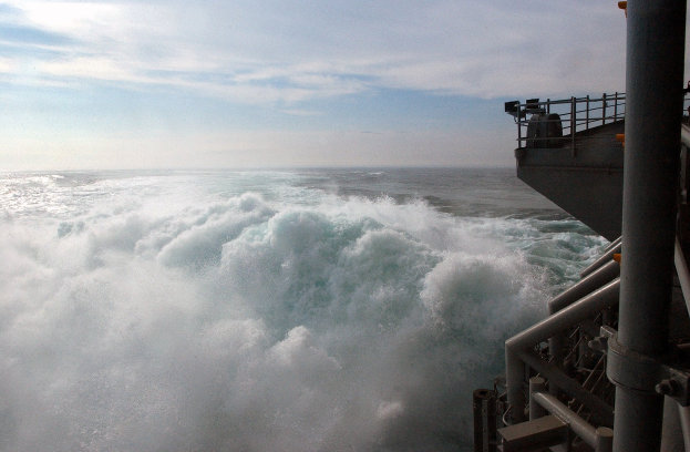 Große Welle bricht über Deck eines Schiffs mit Reling und Pfählen auf der rechten Seite, gegen einen bewölkten Himmel.
