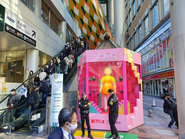 Eine Gruppe von Menschen steht vor einem Gebäude mit Glaswänden, Texttafeln, einige tragen Masken, in der Nähe einer Rolltreppe; ein pinker Kasten in der Mitte zeigt Schaufensterpuppen und trägt "Netflix's new Netflix booth in Tokyo."