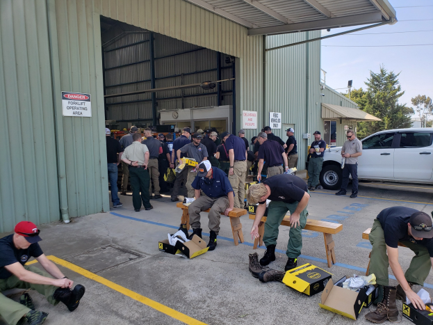 Eine Gruppe von Menschen bei einer Ausbildungssitzung der Nationalgarde, einige sitzen auf Bänken und andere stehen, mit verstreuten Kartons, Schuhen und Gegenständen vor einem Gebäude, Fahrzeugen und Bäumen im Hintergrund unter einem klaren blauen Himmel.