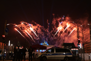 Ein Polizeiwagen steht vor einer Menschenmenge, mit beleuchteten Gebäuden, Bäumen, Polen, Verkehrszeichen, Schildern und Zelten im Hintergrund, unter einem Himmel voller bunter Feuerwerke.