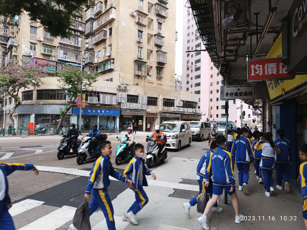 Eine Gruppe von Kindern überquert die Straße vor einem Gebäude, mit Fahrzeugen und Motorrädern auf der Straße, einem Baum links und Gebäuden mit Fenstern, Klimaanlagen und Texttafeln im Hintergrund, mit einem Wasserzeichen unten rechts.