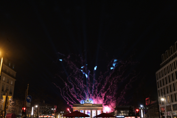 Eine belebte Stadtstraße in Berlin, Deutschland, mit beleuchteten Gebäuden, Polen, Schildern, Verkehrszeichen, Zelten und Fahrzeugen, mit dem Brandenburger Tor im Hintergrund und Feuerwerk am Nachthimmel.
