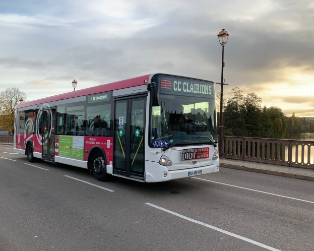 Ein roter und weißer Bus mit der Aufschrift "CC Clairions" fährt an einem Wasserfrontweg mit einem Geländer auf einer Seite und Laternen auf der anderen, Bäume und Gebäude im Hintergrund unter einem bewölkten Himmel.