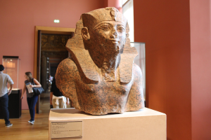 Ein Büste eines ägyptischen Pharaos in einem Museum mit einer Texttafel, einer Gruppe von Menschen, einer Frau mit einer Tasche, einer Tür, einer Wand und einer Decke mit Lampen.