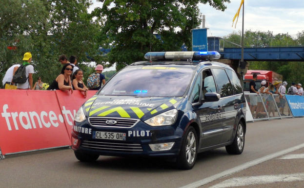 Ein Polizeiwagen fährt eine Straße entlang neben einer Menge, wobei einige Menschen Mützen tragen und Taschen tragen, ein Banner auf der linken Seite, Geländer mit Bannern dahinter und ein Hintergrund mit Bäumen, einer Brücke, einer Fahne auf einem Mast und einem bewölkten Himmel.