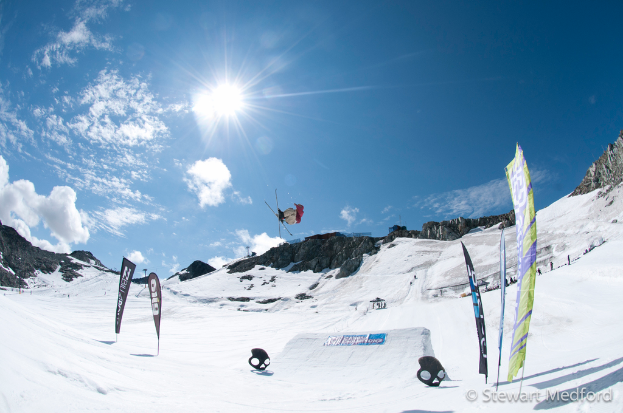 Eine Person in Skier ist in der Luft mit schneebedeckten Bergen im Hintergrund.