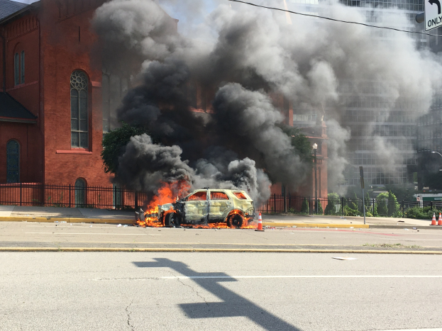 Ein brennendes Auto am Straßenrand mit Rauchwolken, umgeben von Gebäuden, Bäumen und anderen städtischen Elementen unter einem klaren Himmel.