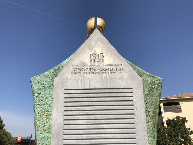 Ein steinernes Denkmal in einem Park mit einer Uhr darauf, umgeben von Bäumen und Gebäuden, mit einer Inschrift, die den 24. Jahrestag des Völkermords in Armenien erinnert.