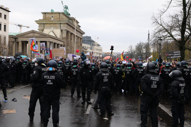 Eine große Gruppe von Polizisten steht vor einer Menge mit Schildern, Luftballons und Helmen, mit Gebäuden, Bäumen und einem bewölkten Himmel im Hintergrund.