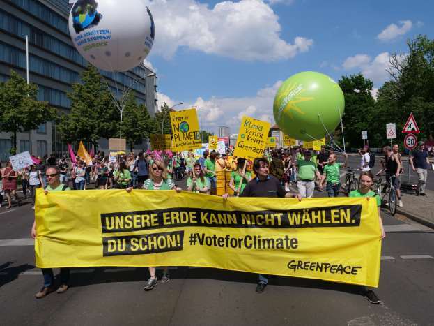 Eine Gruppe von Menschen geht eine Straße entlang und hält ein gelbes Banner mit der Aufschrift "Wähle für das Klima", mit Ballons, Schildern, Fahrrädern, Bäumen, Laternenmasten und Gebäuden im Hintergrund unter einem klaren blauen Himmel.