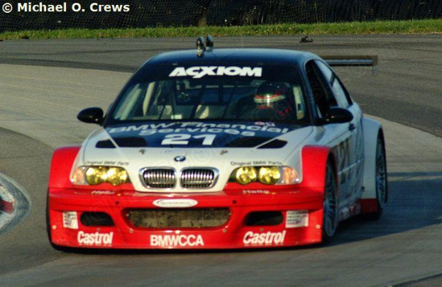 Eine Person mit Helm fährt einen BMW M3 GT3 Rennwagen auf einer von Gras und einem Maschendrahtzaun umgebenen Strecke, mit einem Wasserzeichen in der linken oberen Ecke.