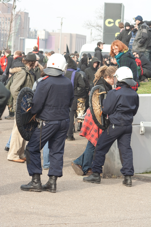 Eine Gruppe von Menschen, die auf einer Straße gehen, mit zwei Personen vorne, die wie Polizei aussehen, Gebäude im Hintergrund und Boden unten.