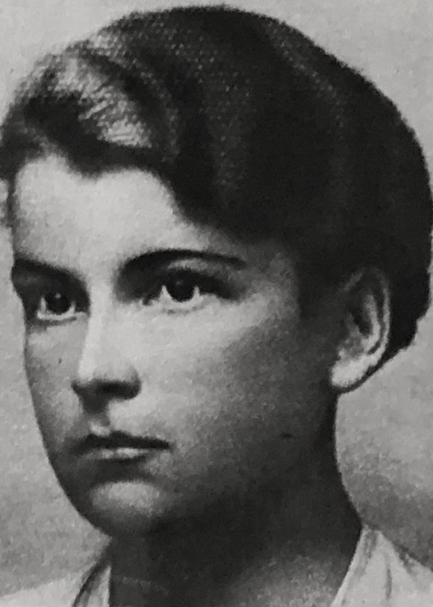 Ein Schwarz-Weiß-Foto einer Frau mit kurzen, zurückgekämmten Haaren, die ein weißes Hemd trägt, mit ernster Miene, fokussierten Augen und leicht geöffneten Lippen, die Hände vor der Brust verschränkt.