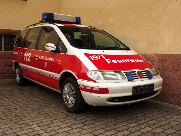 Rote und weiße Ambulanz mit Text und Zahlen vor einem Fensterbau geparkt.