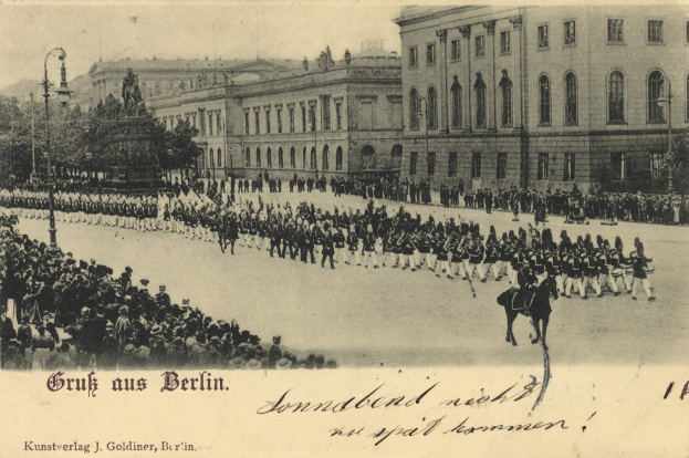 Ein altes Schwarz-Weiß-Foto eines Umzugs in Berlin, Deutschland, mit Menschen auf dem Boden und Pferden, einer Statue, Straßenlaternen, Bäumen, Gebäuden mit Fenstern und einem bewölkten Himmel, mit Text am unteren Rand.