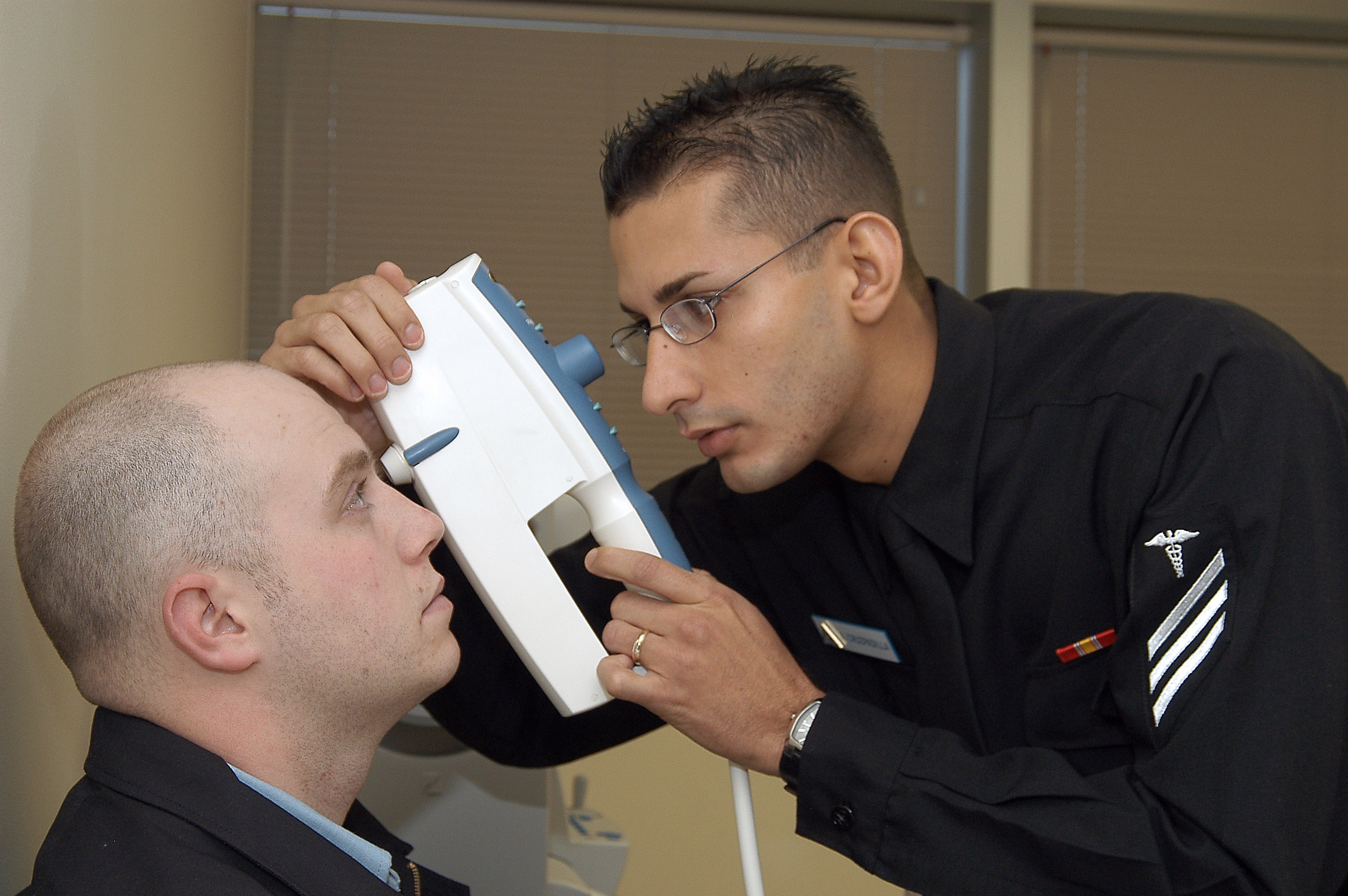 Ein Mann in einem schwarzen Hemd und Krawatte bei einer Augenscreening-Untersuchung durch einen Arzt in einer Brille, der ein Objekt hält, mit Jalousien im Hintergrund.