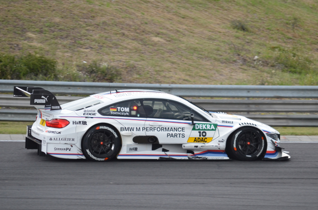 Ein BMW M4 DTM-Rennwagen fährt auf einer Rennstrecke, umgeben von saftig grünem Gras, mit einer Absperrung im Hintergrund.