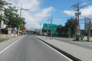 Arterielle Straße mit Fahrzeugen, Strommasten, Schildern, Gebäuden, Bäumen und einem bewölkten Himmel im Hintergrund.