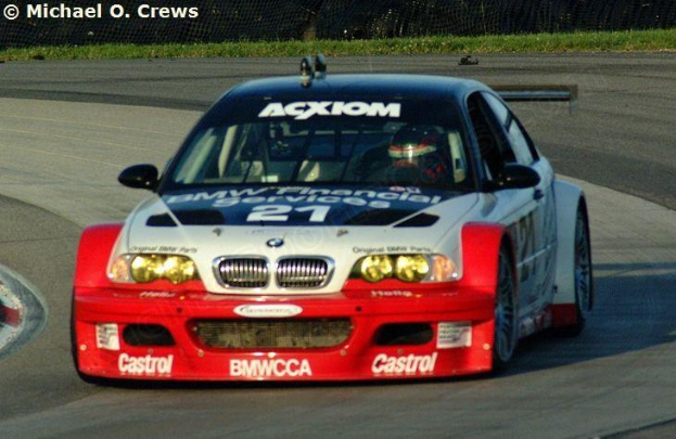 Eine Person in einem Helm fährt einen BMW M3 GT3 Rennwagen auf einer von Gras und einem Maschendrahtzaun umgebenen Strecke, mit sichtbarem Text, Zahlen und einem Wasserzeichen in der linken oberen Ecke.